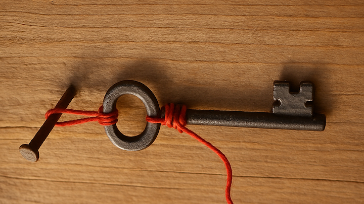 Ein alter Schlüssel hängt seitlich an einem roten Faden, der um seinen Ring und einen schräg eingeschlagenen Nagel gewickelt ist. Das Bild ist um 90 Grad gedreht – der Schlüssel wirkt wie angehalten in einem Moment zwischen Festhalten und Loslassen.